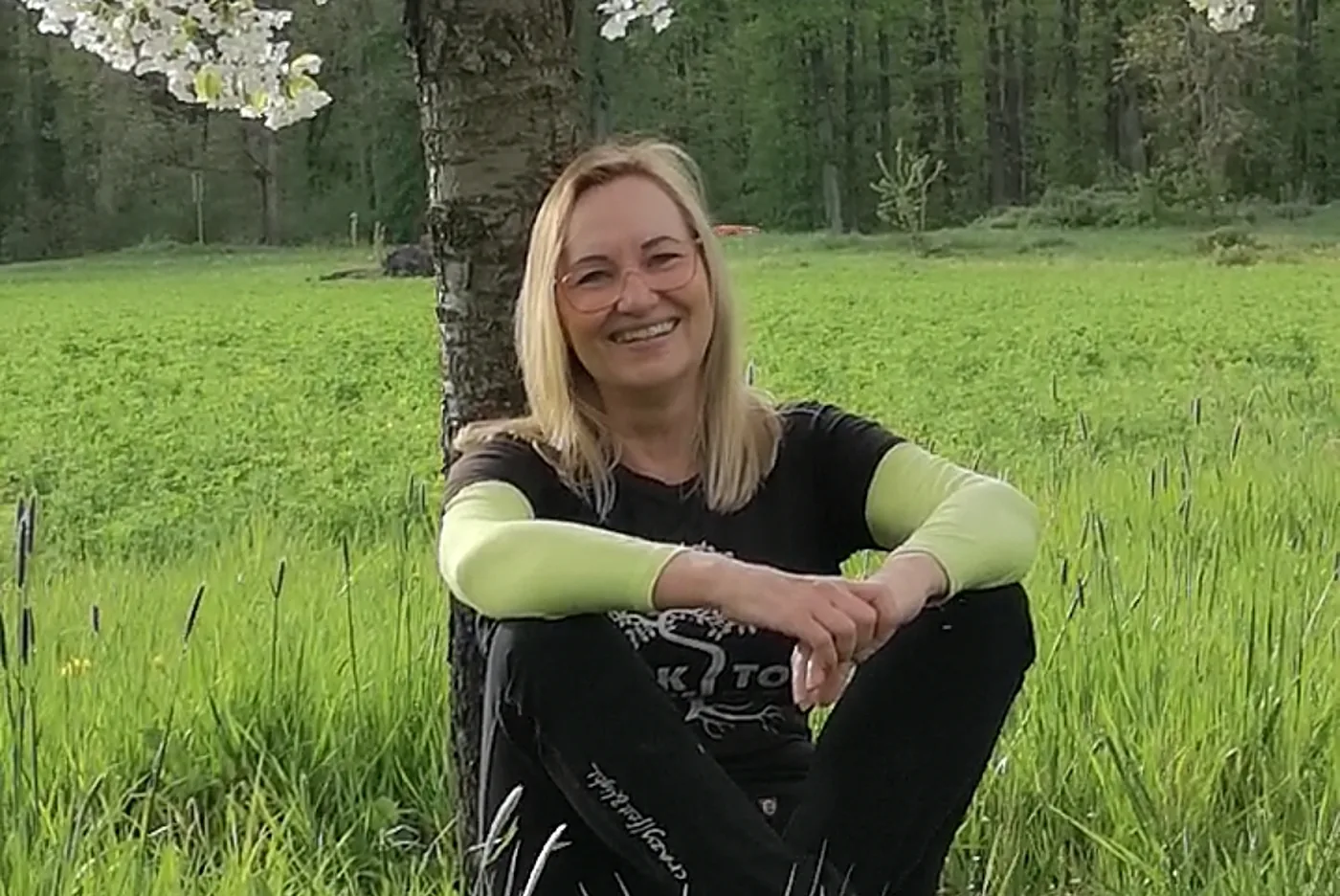 Sabine Rößler sitzt entspannt und lächelnd unter einem blühenden Kirschbaum auf einer grünen Wiese