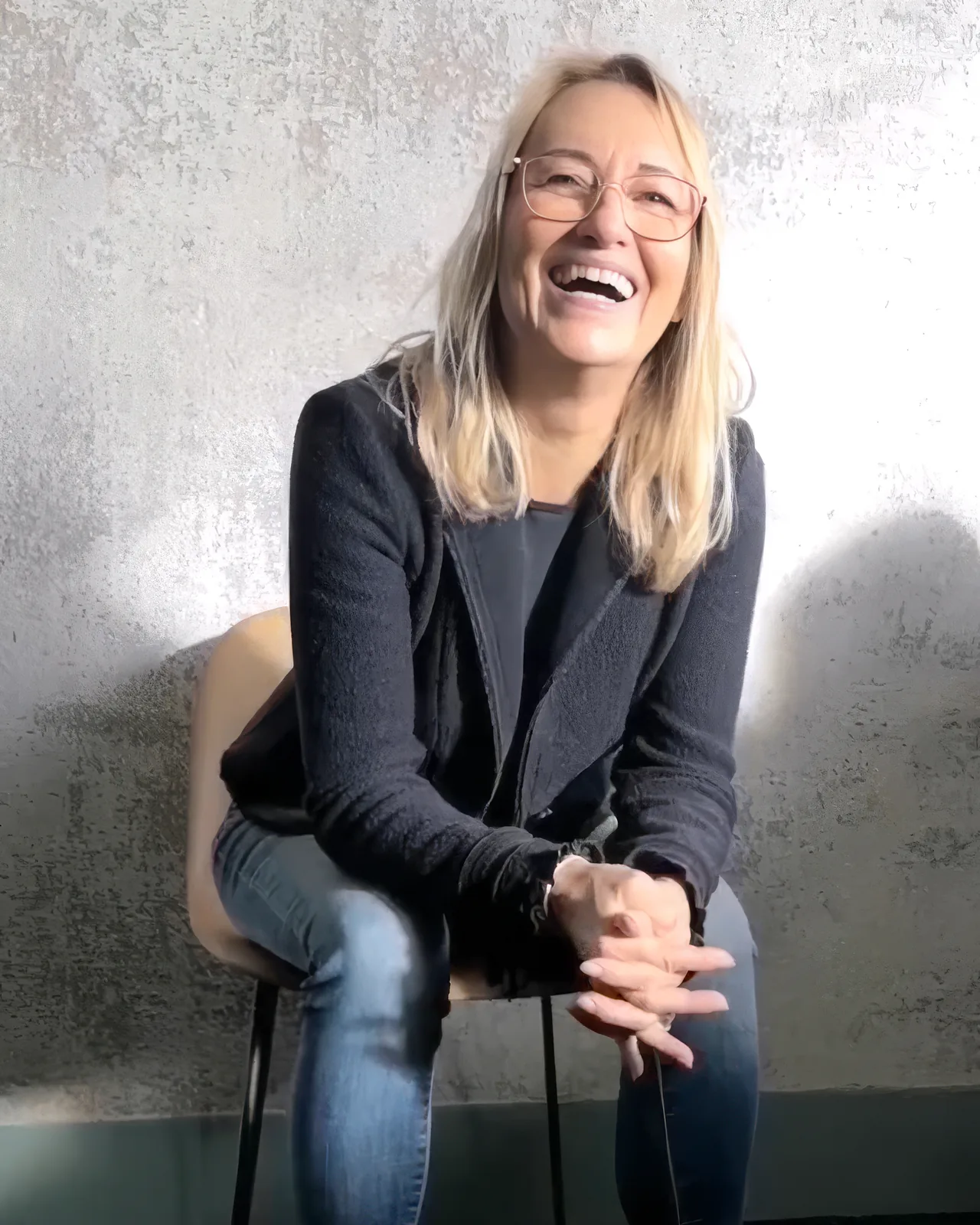 Sabine Rößler sitzt lachend auf einem Hocker vor einer hellen Wand