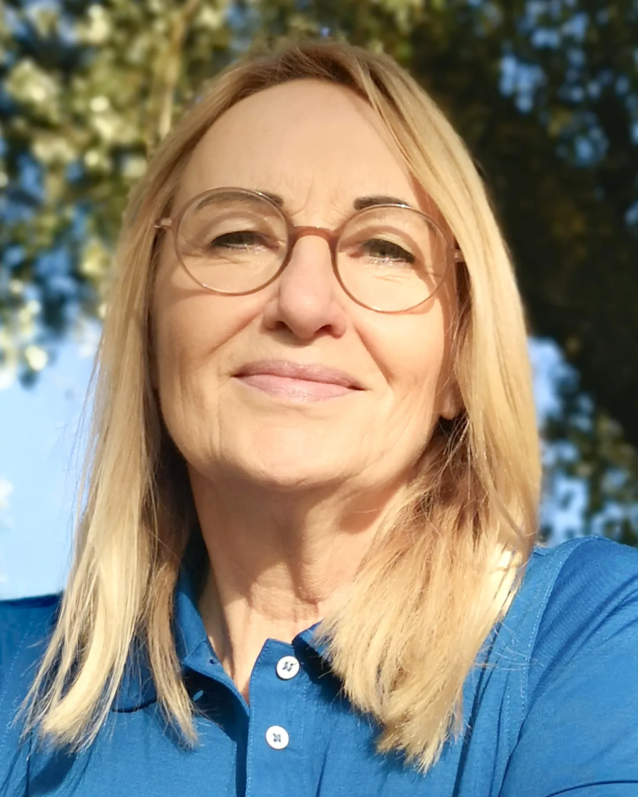 Sabine Rößler im Freien, lächelnd, im Hintergrund Baumkronen und blauer Himmel